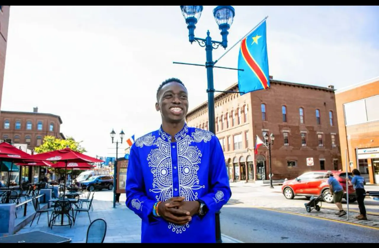 Fiston smiling on a downtown Concord street in an embroidered blue shirt, with the flag of the DR Congo behind him.